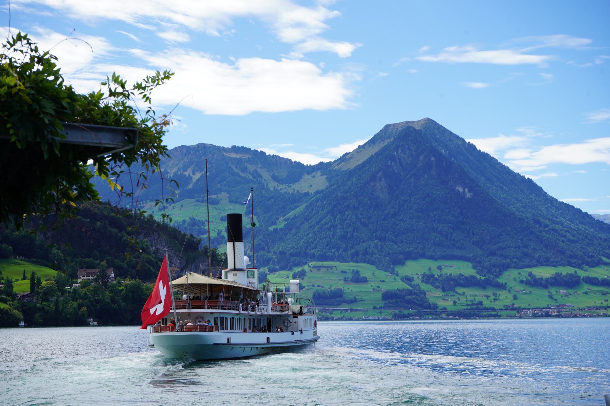A paddle steamer flying the Swiss flag leaving the shore of Lake Lucerne, a pyramid of a mountain behind