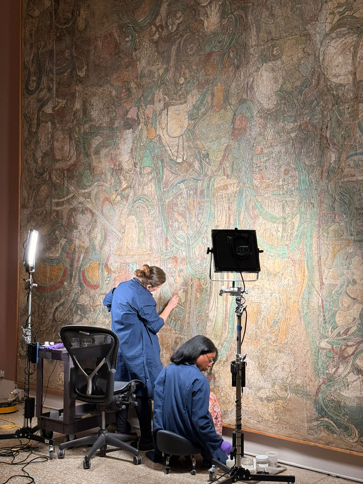 Two conservators in blue coats working under lamps on an enormous faded Buddhist wall mural at the Met